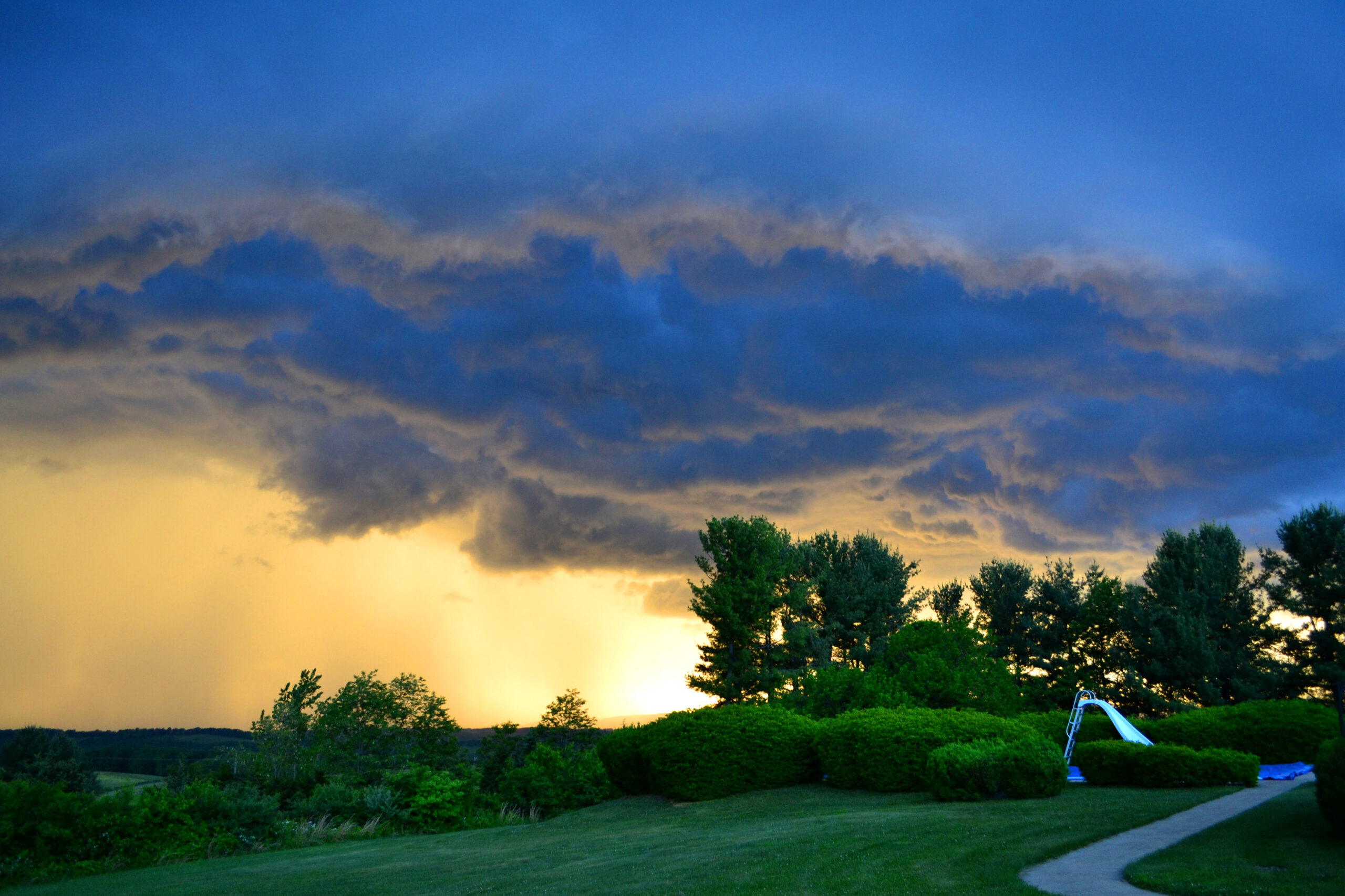 Dramatic sunset sky with dark thunderstorm clouds and vibrant orange light, framed by green trees and a grassy landscape, reflecting the transformative energy of ORG NYC's organizing services.