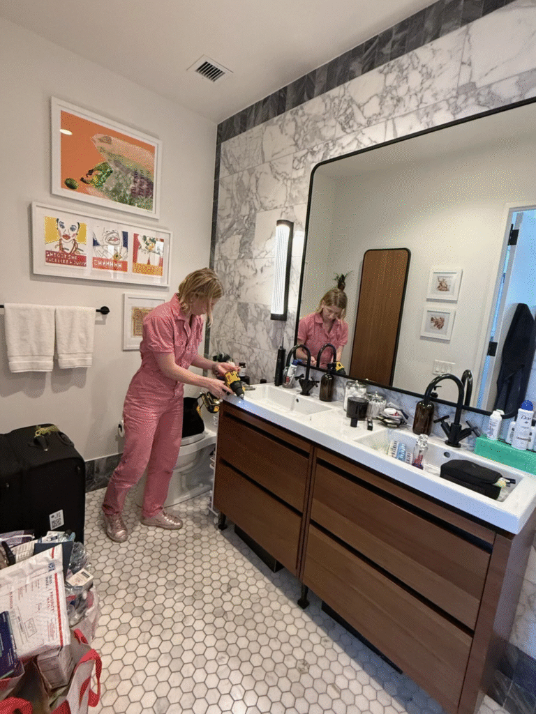 Woman in pink jumpsuit organizing a bathroom space with a drill, featuring a modern vanity, marble walls, and various personal items scattered around, illustrating practical home organization.