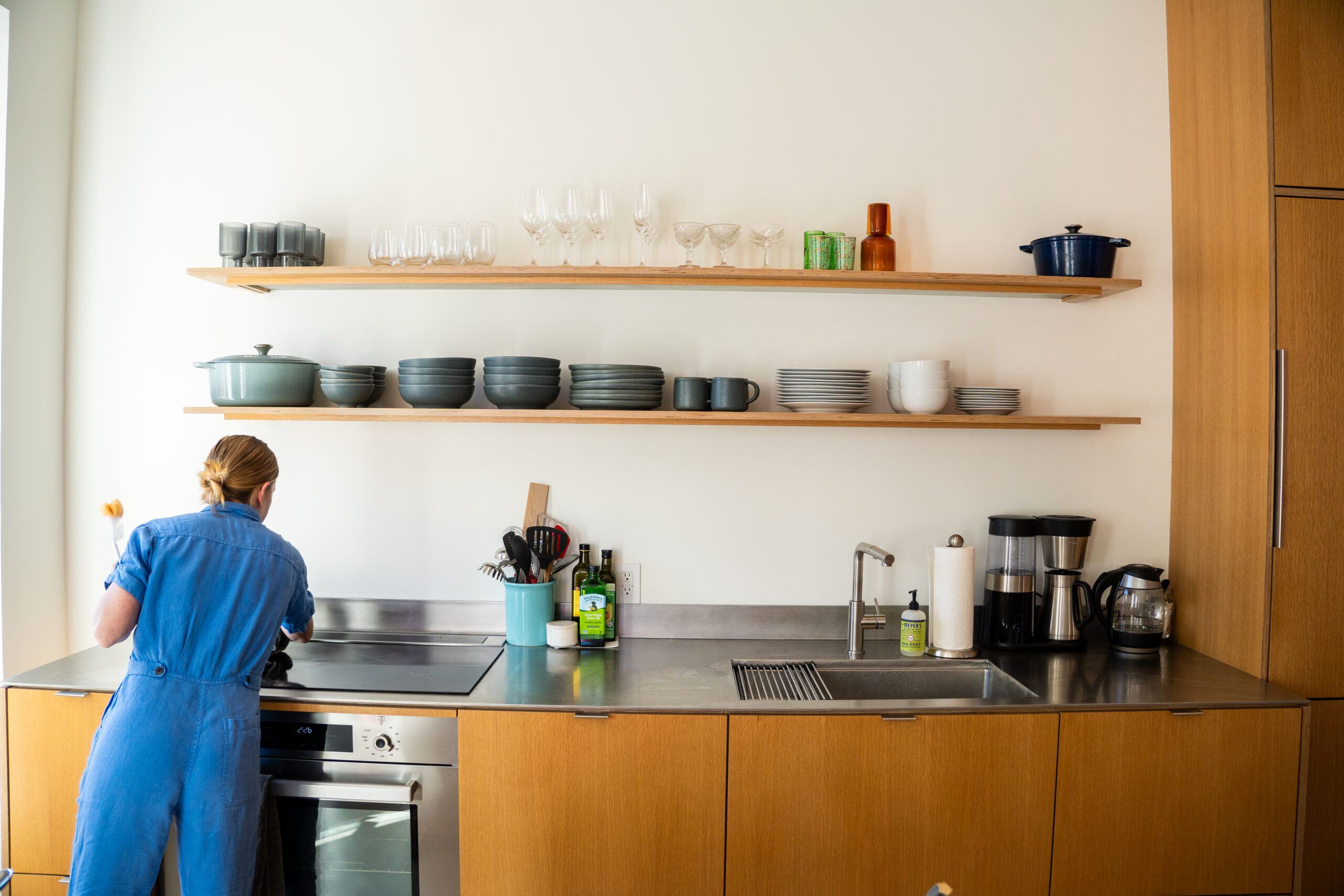 Woman in blue outfit cleaning a modern kitchen countertop with organized shelves displaying various dishes and glassware, emphasizing kitchen clarity and functionality.