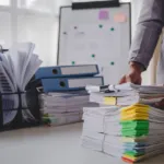 A person organizing piles of documents for an office paperwork organization system with binders on a bright workspace desk
