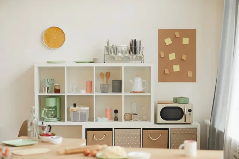 Bright kitchen with a white cube shelf holding dishes, a coffee maker, a microwave and woven storage bins below a cork board of sticky notes