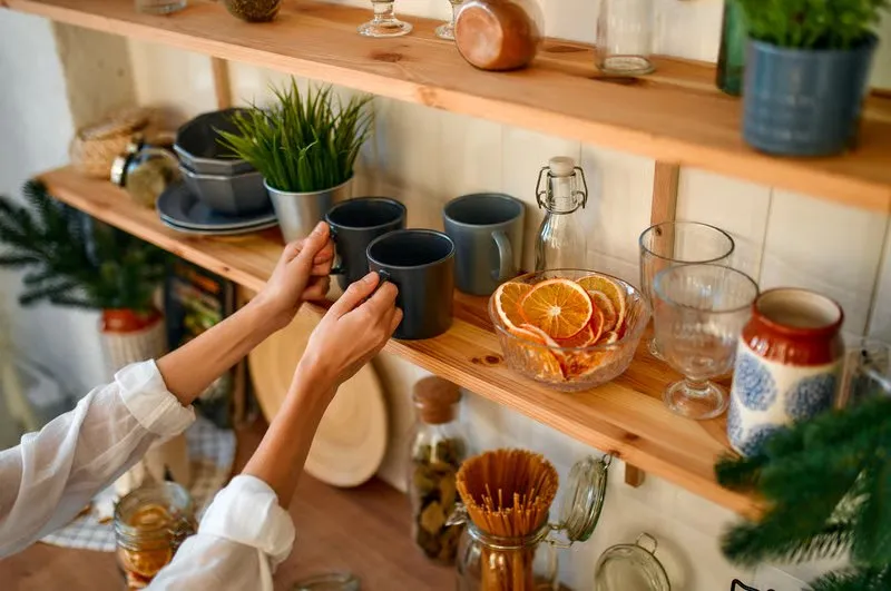 Hands arranging gray mugs on open wooden kitchen shelves beside glassware and a bowl of dried oranges